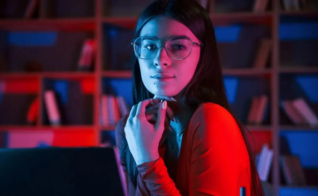 Beautiful woman in glasses and red wear is sitting by the laptop in dark room with neon lighting